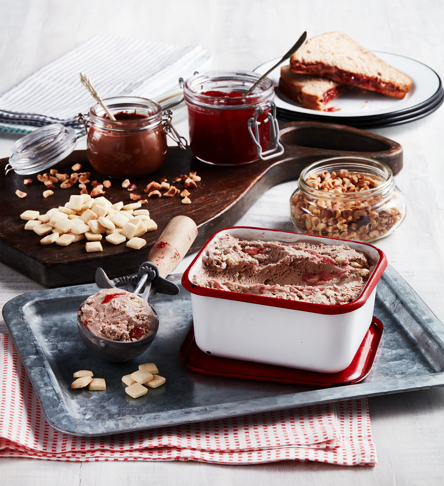 A variety of toppings and ingredients for making ice cream, including jars of jam, chopped nuts, marshmallows, and a container of ice cream with a spoon, on a table with a serving tray and a slice of strawberry jam sandwich.