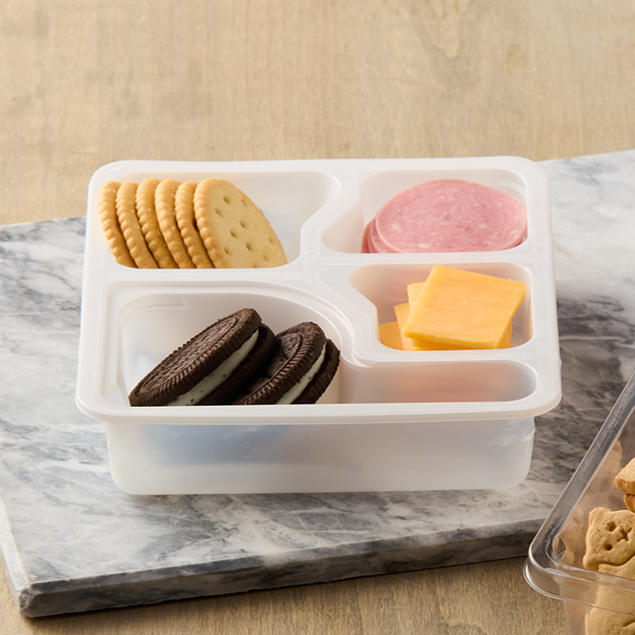 A white plastic tray with sections holding round crackers, sliced ham, sliced cheddar cheese, and Oreo cookies, placed on a marble surface.