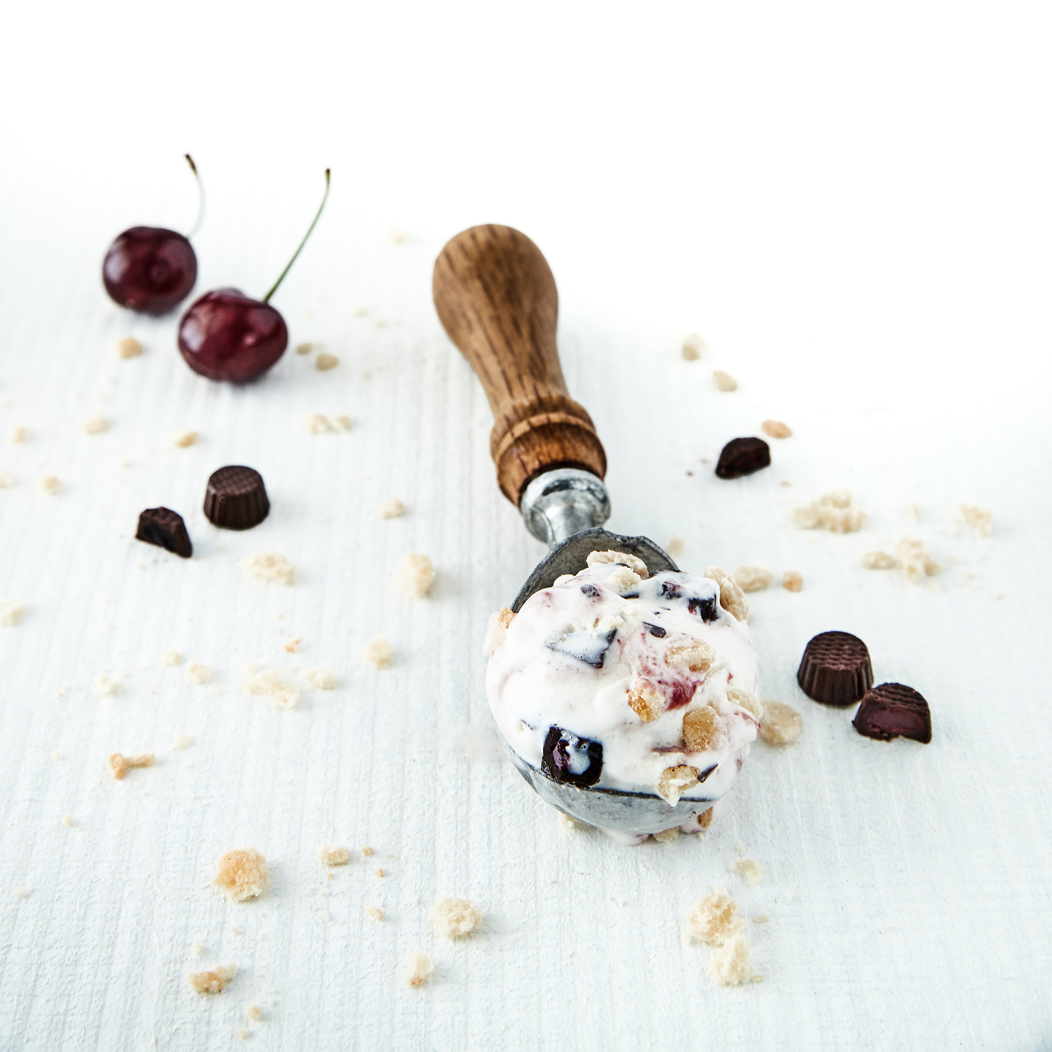 Scooping cherry and chocolate chip ice cream with cherries and chocolate pieces scattered on a white surface.