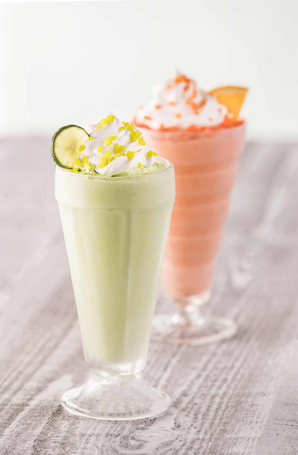 Two milkshakes, one green with lime and whipped cream, one pink with whipped cream and orange sprinkles, on a wooden surface.