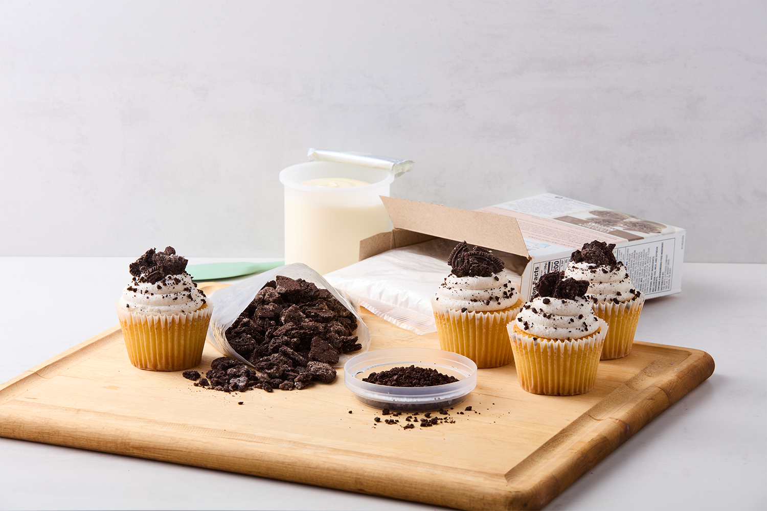Four vanilla cupcakes with cookies and cream frosting and crushed cookies on top, placed on a wooden cutting board, with an open cookie crumble bag, a container of cookie crumbs, a container of Oreos, a box of milk, and a spoon in the background.