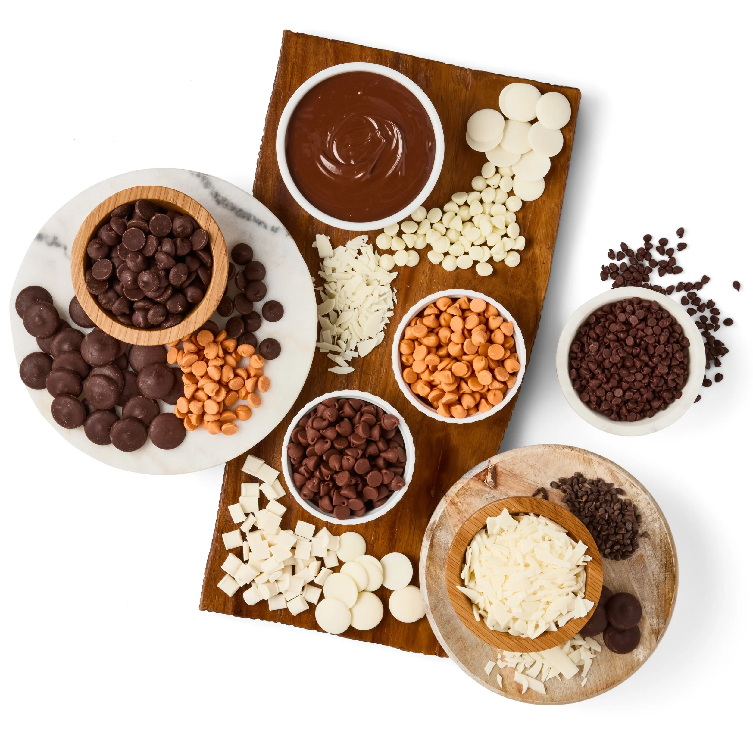 Assorted chocolate chips and chocolate melts in bowls and on a wooden and marble surface.