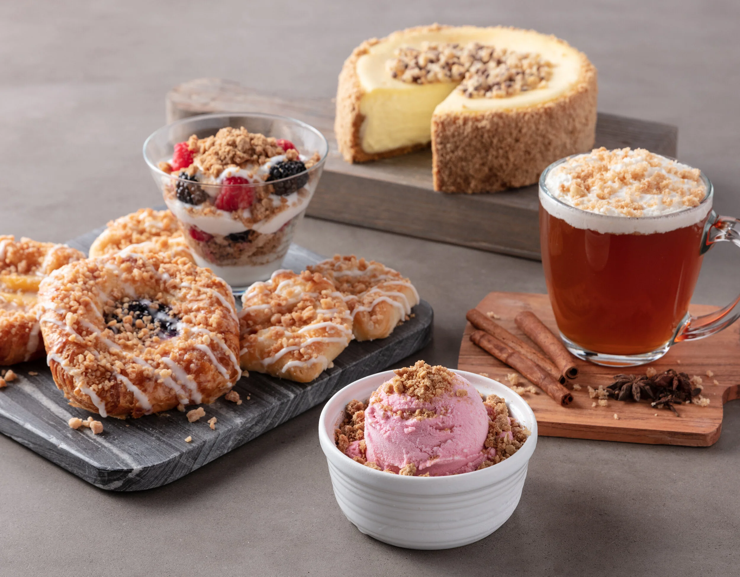 Assorted desserts including berry cheesecake, mixed berry parfait, cookies and cream ice cream, blueberry muffins, and a tea with cinnamon sticks on a wooden board and slate serving platters