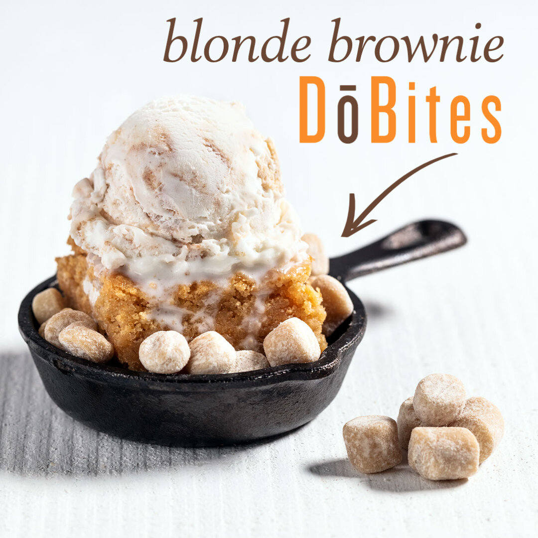 A small cast iron skillet containing a scoop of blonde brownie ice cream with cookie dough pieces surrounding it, with text indicating 'blonde brownie DōBites' and an arrow pointing to the ice cream.