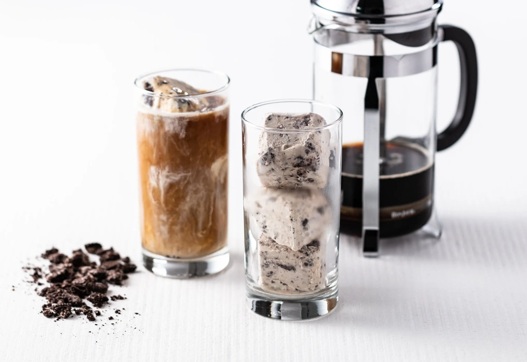 Two glasses of iced coffee with ice cubes next to a French press coffee maker on a white surface with coffee grounds nearby.