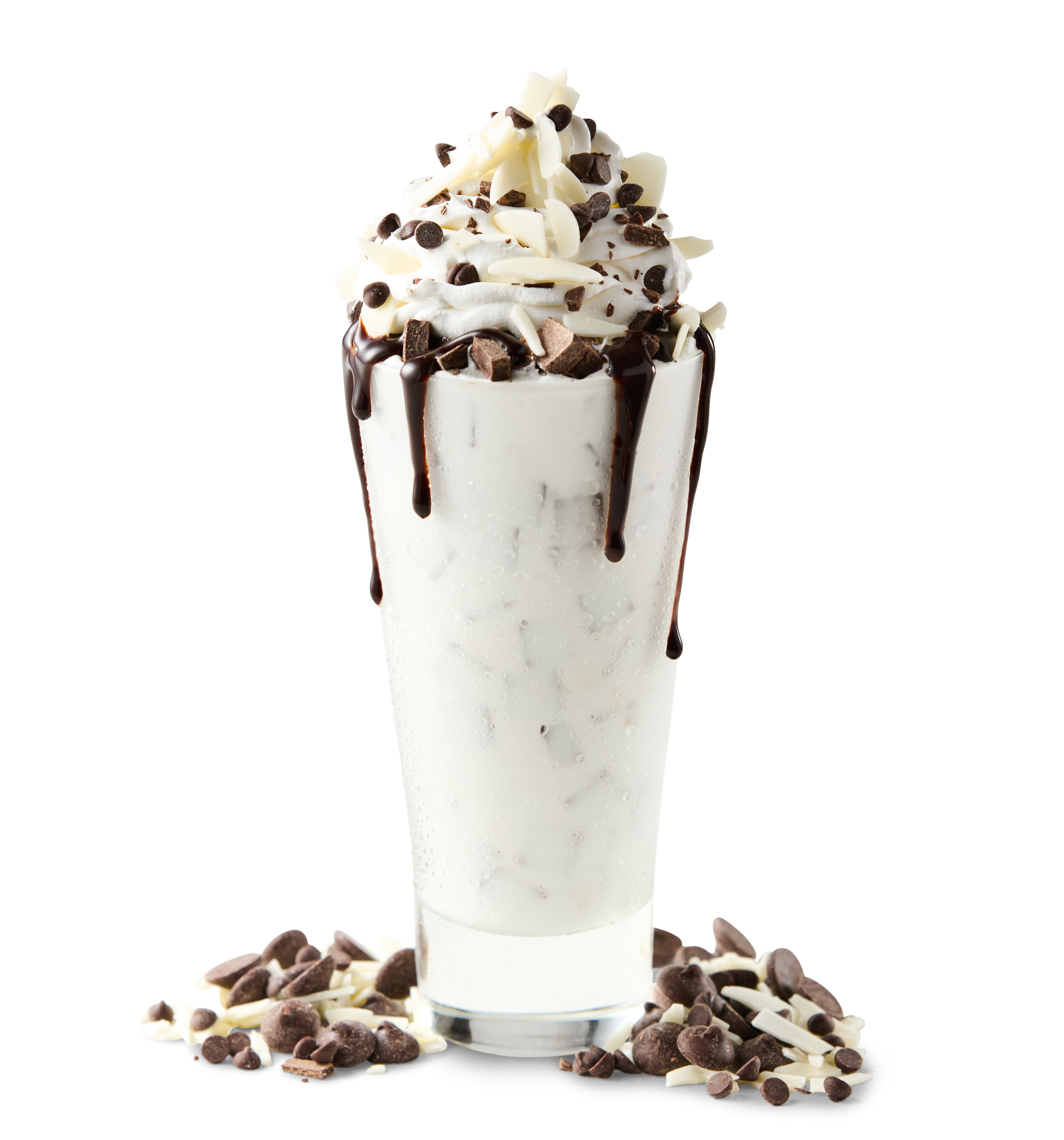 A tall glass of vanilla milkshake topped with whipped cream, chocolate shavings, and chocolate chips, with some chocolate chips and shavings spilled on the surface beneath.
