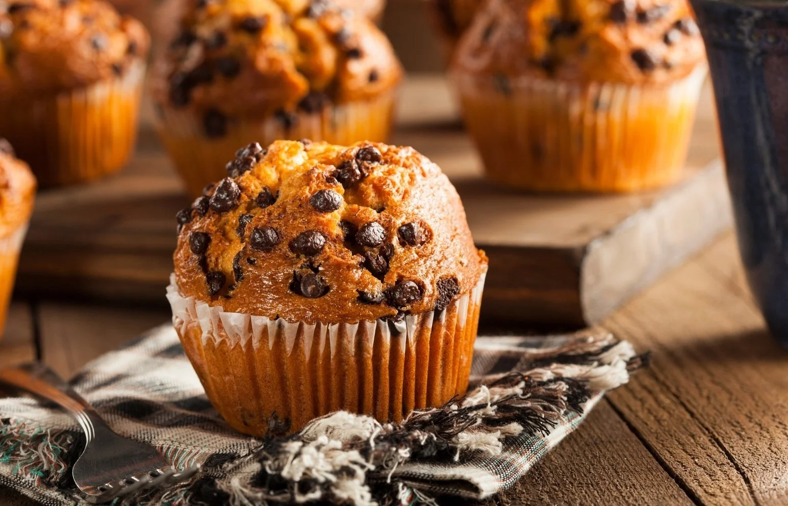 Chocolate chip muffin on a cloth napkin with additional muffins in the background on a wooden surface.