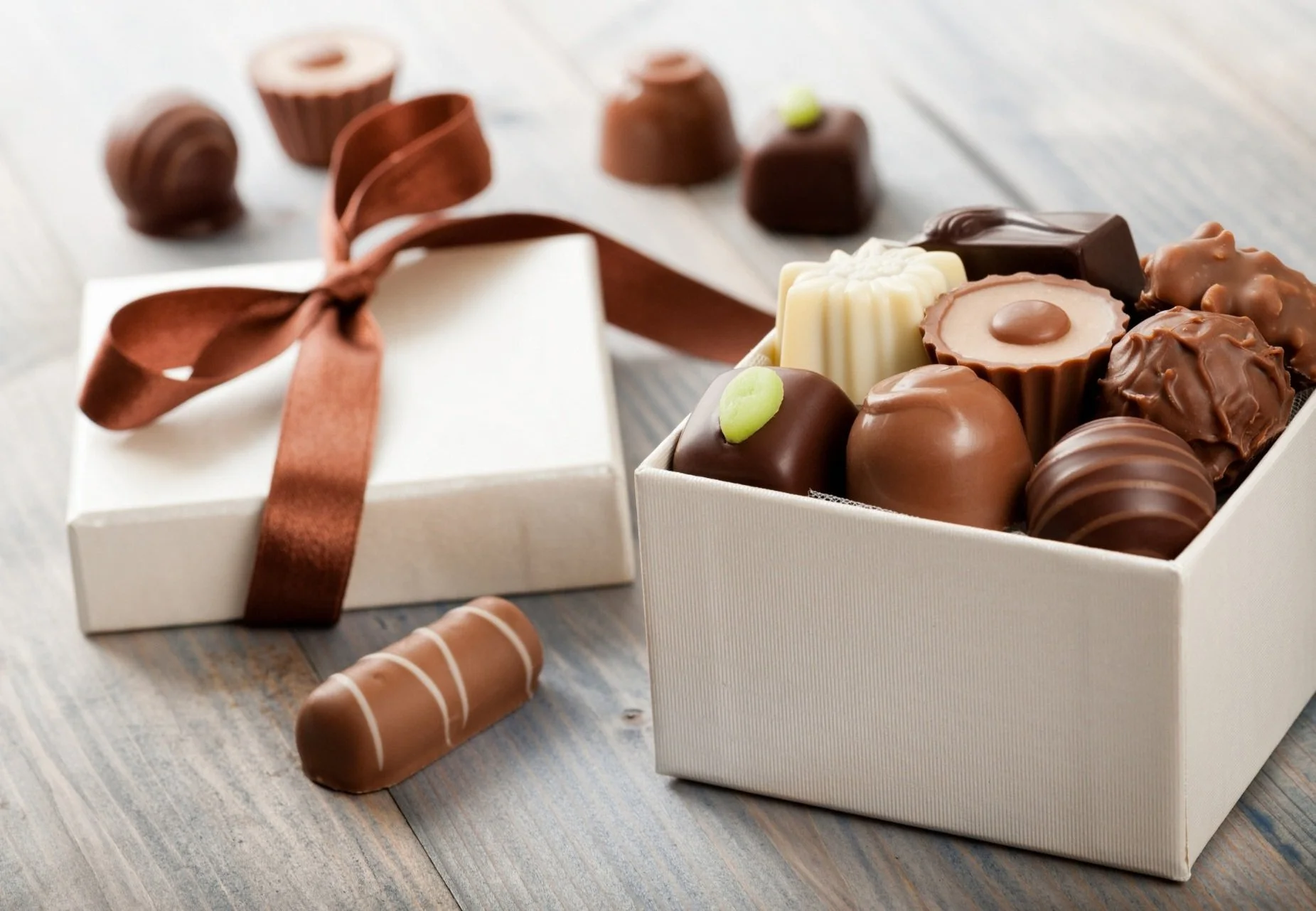 A white gift box with a brown ribbon, open to reveal assorted chocolates, with more chocolates placed outside on a wooden surface
