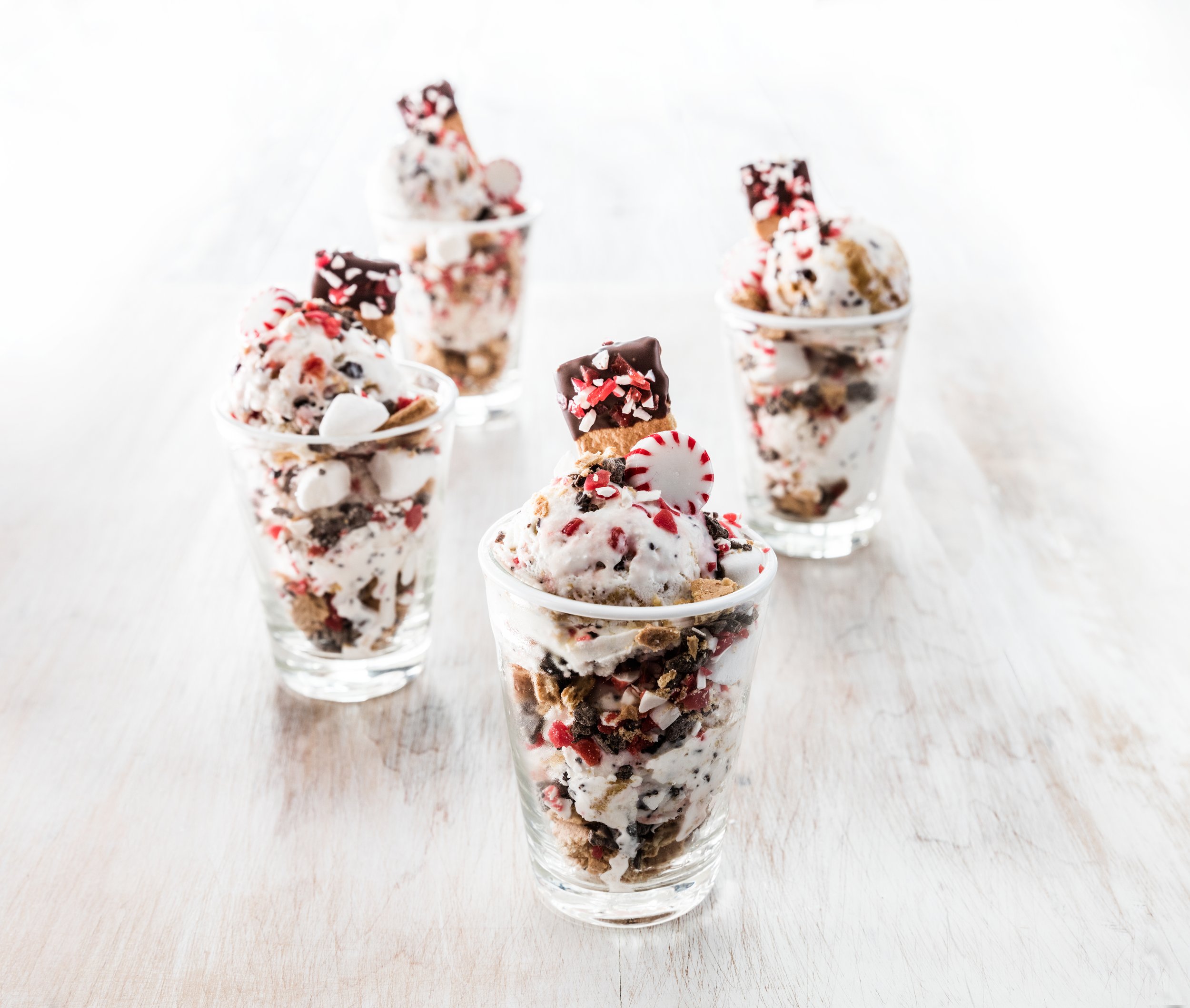 Four glasses filled with ice cream topped with chocolate, peppermint, and crushed candies, set on a light wooden surface.