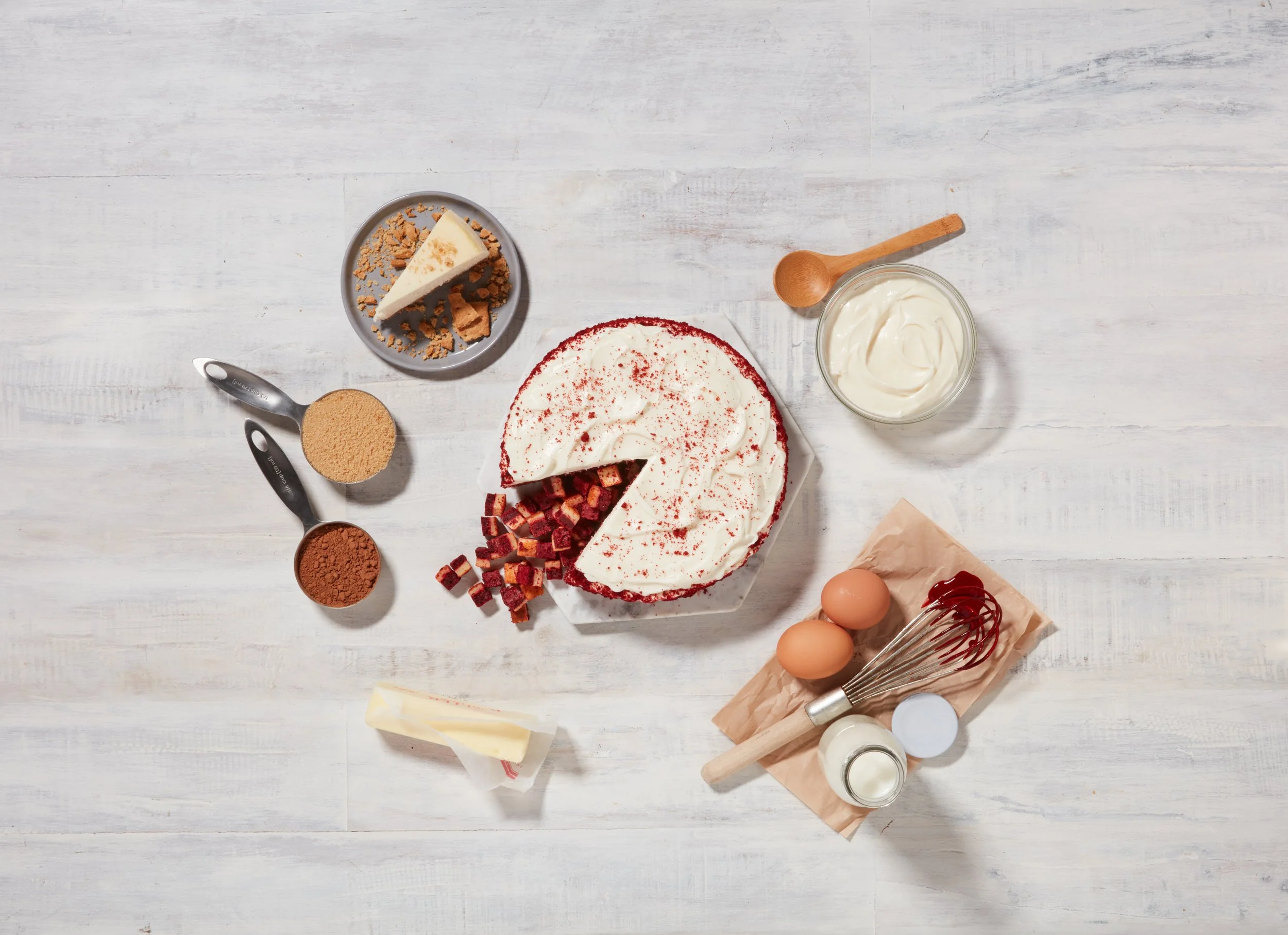 Unfinished birthday cake with white frosting and red sprinkles, surrounded by baking ingredients and tools on a white wooden surface.