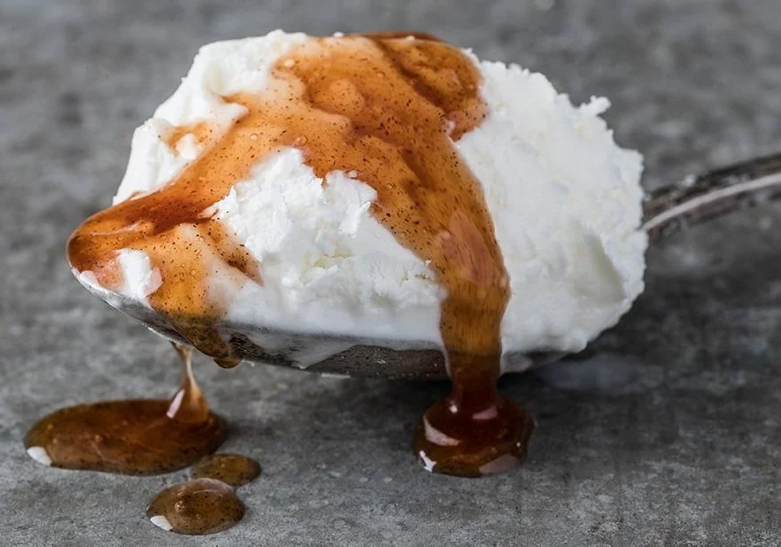 Spoonful of vanilla ice cream with caramel sauce drizzling over it, on a gray surface.