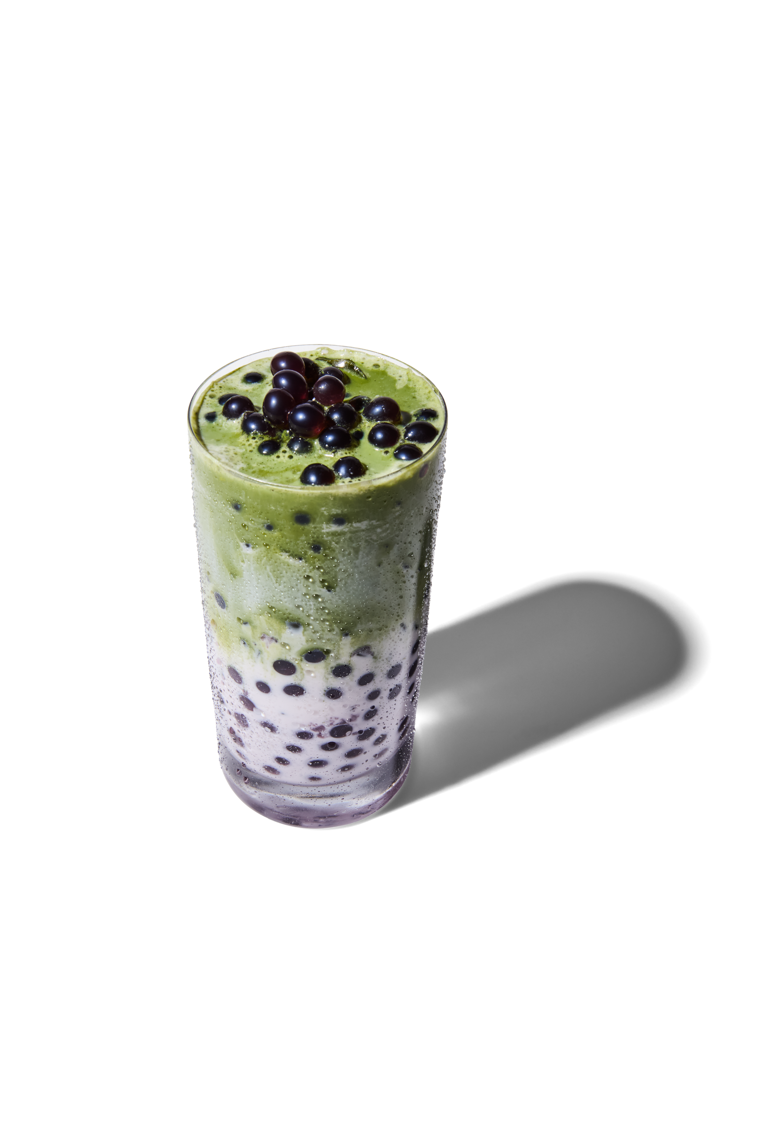A tall glass of bubble tea with black tapioca pearls, topped with green matcha foam and black tapioca pearls.