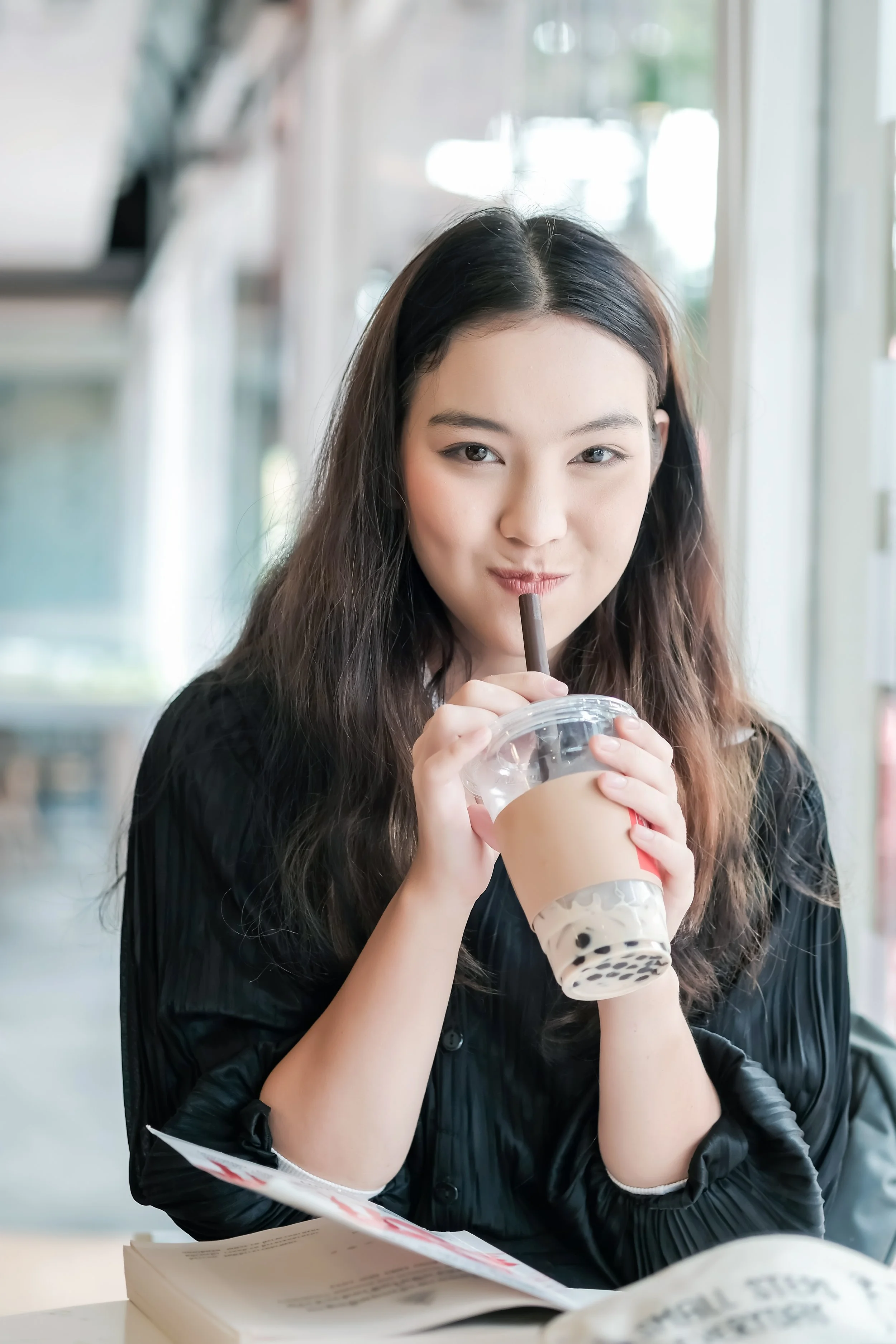 A young woman with long dark hair, wearing a black top, enjoys a drink at a cafe, holding a plastic cup with a brown sleeve and a black straw, with a book and papers in front of her.