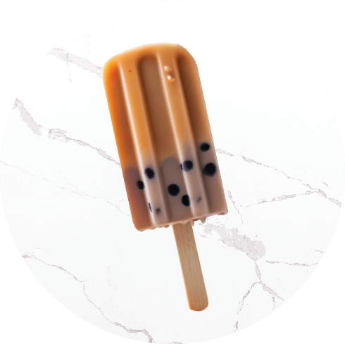 Brown bubble tea-shaped popsicle with black tapioca pearls on a wooden stick, placed on a white marble surface.