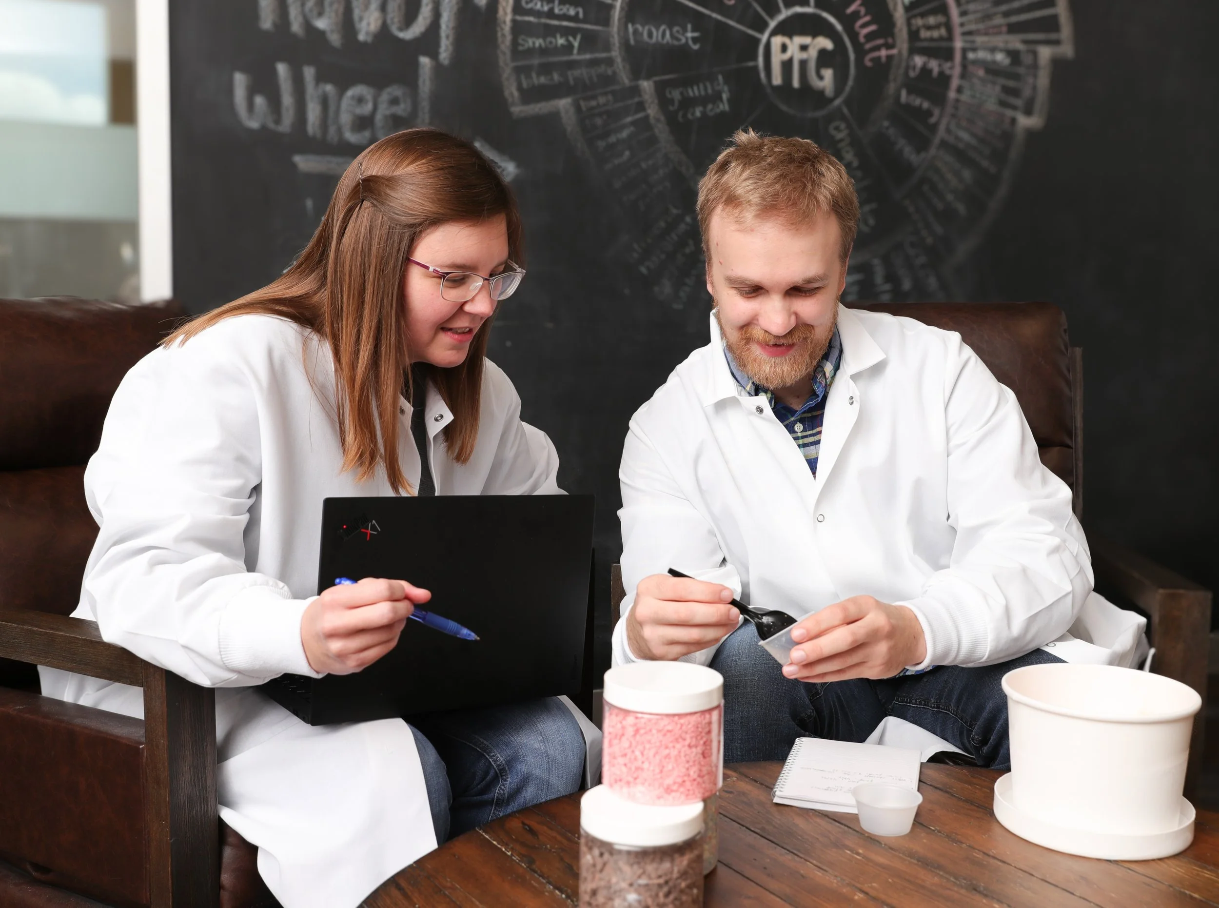 Two scientists, a woman and a man, in white lab coats working together on a project in a laboratory. The woman is holding a pen and a laptop, while the man is using a spatula and a small container. There are various containers and notes on the wooden table, and a blackboard with scientific diagrams in the background.