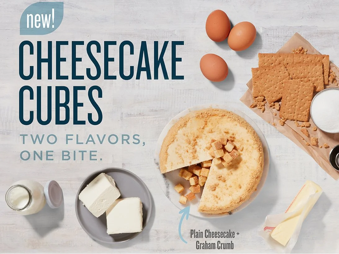 Display of new cheesecake cubes in two flavors, plain cheesecake and graham crumb, with eggs, graham crackers, milk, and butter ingredients on a white wooden surface.