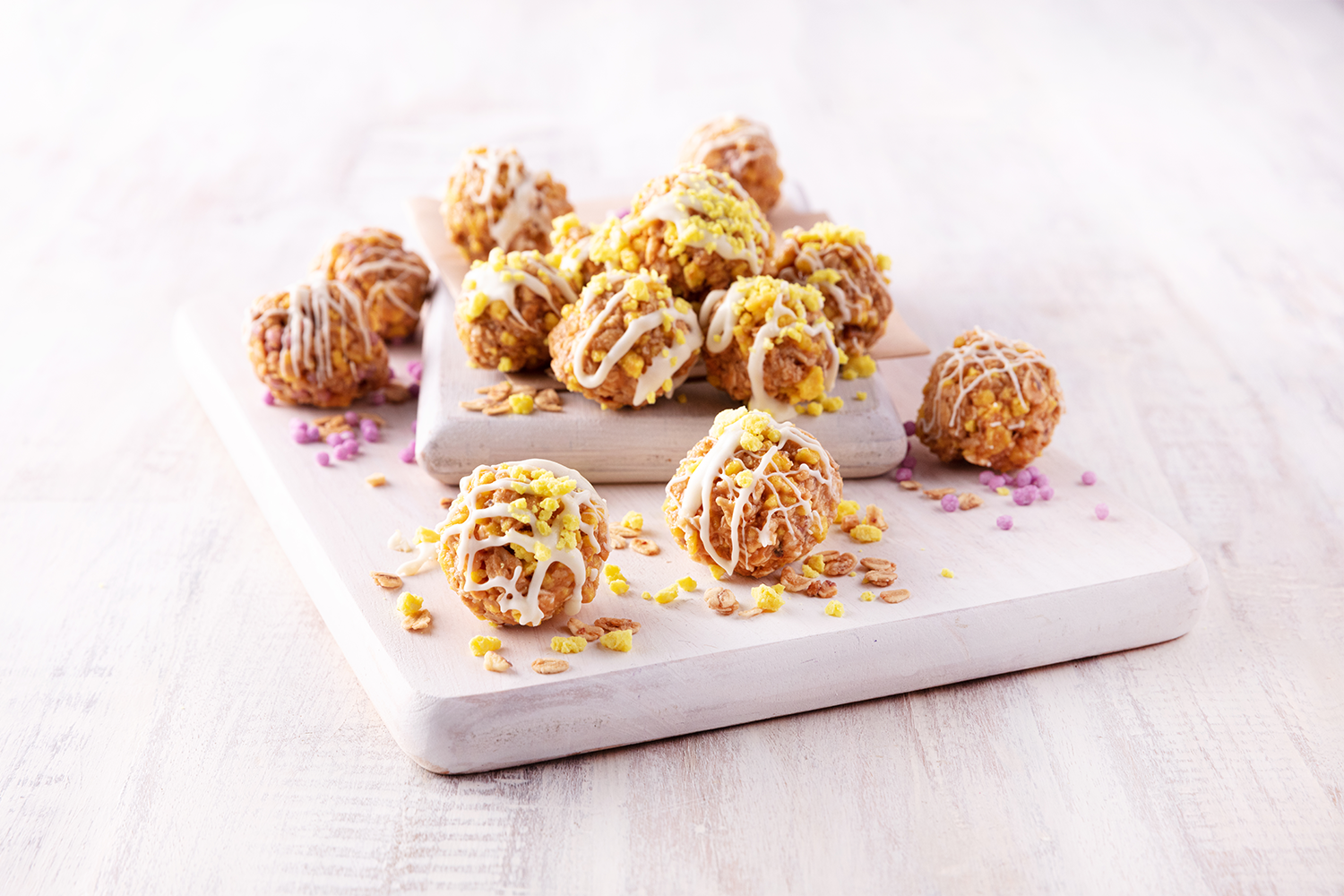 Plate of assorted cookie balls with yellow and pink sprinkles and white drizzle on a white wooden table.