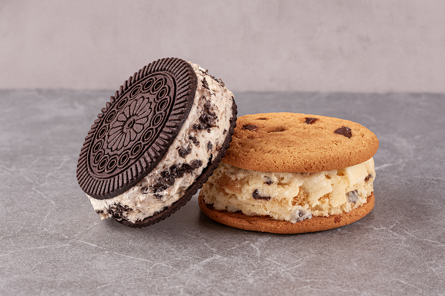 Close-up of an ice cream sandwich with cookies and cream ice cream and an Oreo cookie, next to a chocolate chip cookie with cream filling and ice cream and chocolate chips on top.