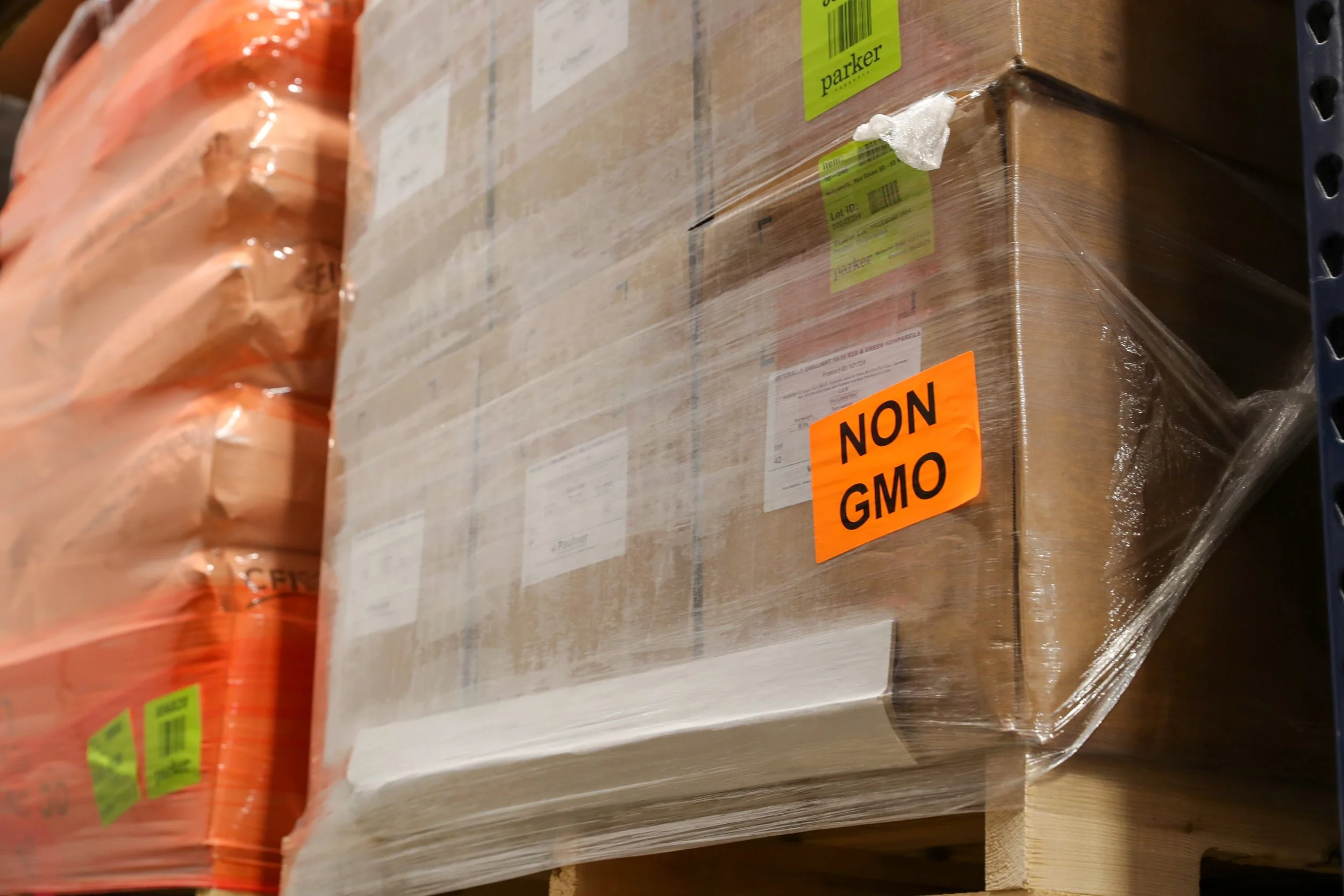 A cardboard box on a warehouse shelf with an orange label that reads 'NON GMO'. The box is wrapped in clear plastic and surrounded by other wrapped packages.