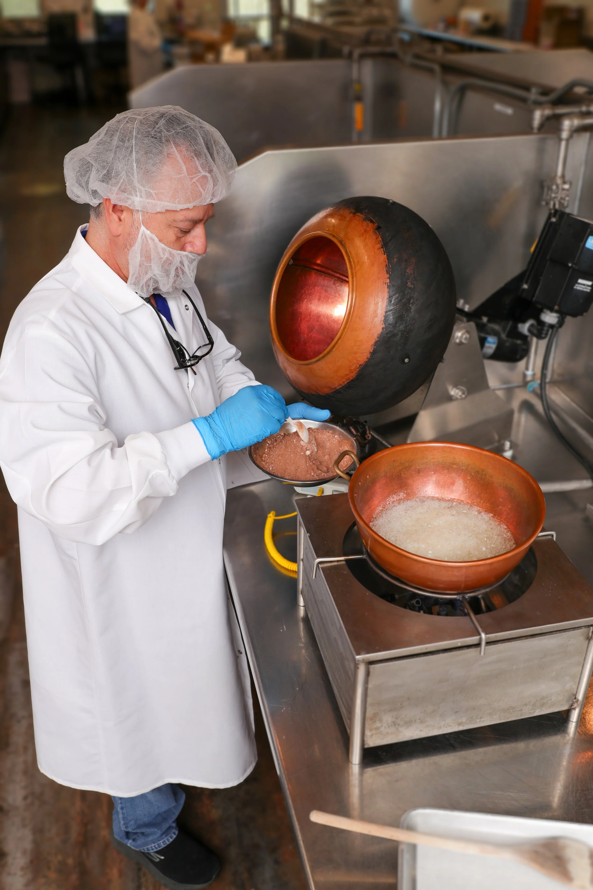 A man wearing a white lab coat, a hairnet, and blue gloves is working with a large, industrial melting machine and cast iron cookware, stirring a mixture inside the cookware.