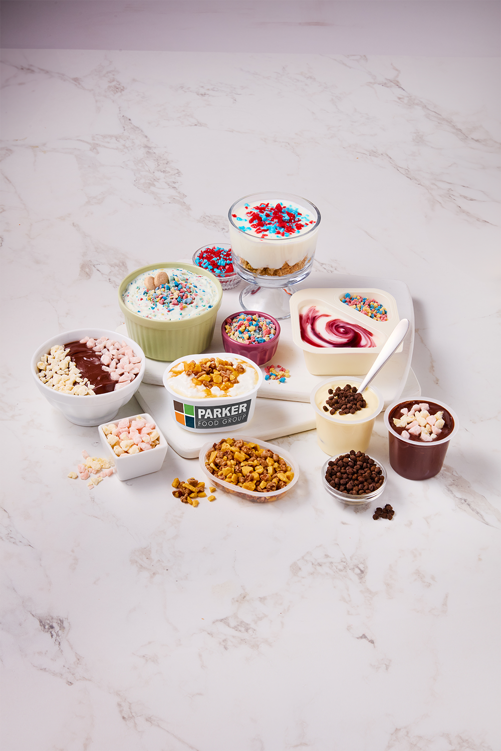 Assorted desserts and toppings including ice cream, yogurt, chocolate pudding, cereal, sprinkles, and whipped toppings arranged on a marble surface.