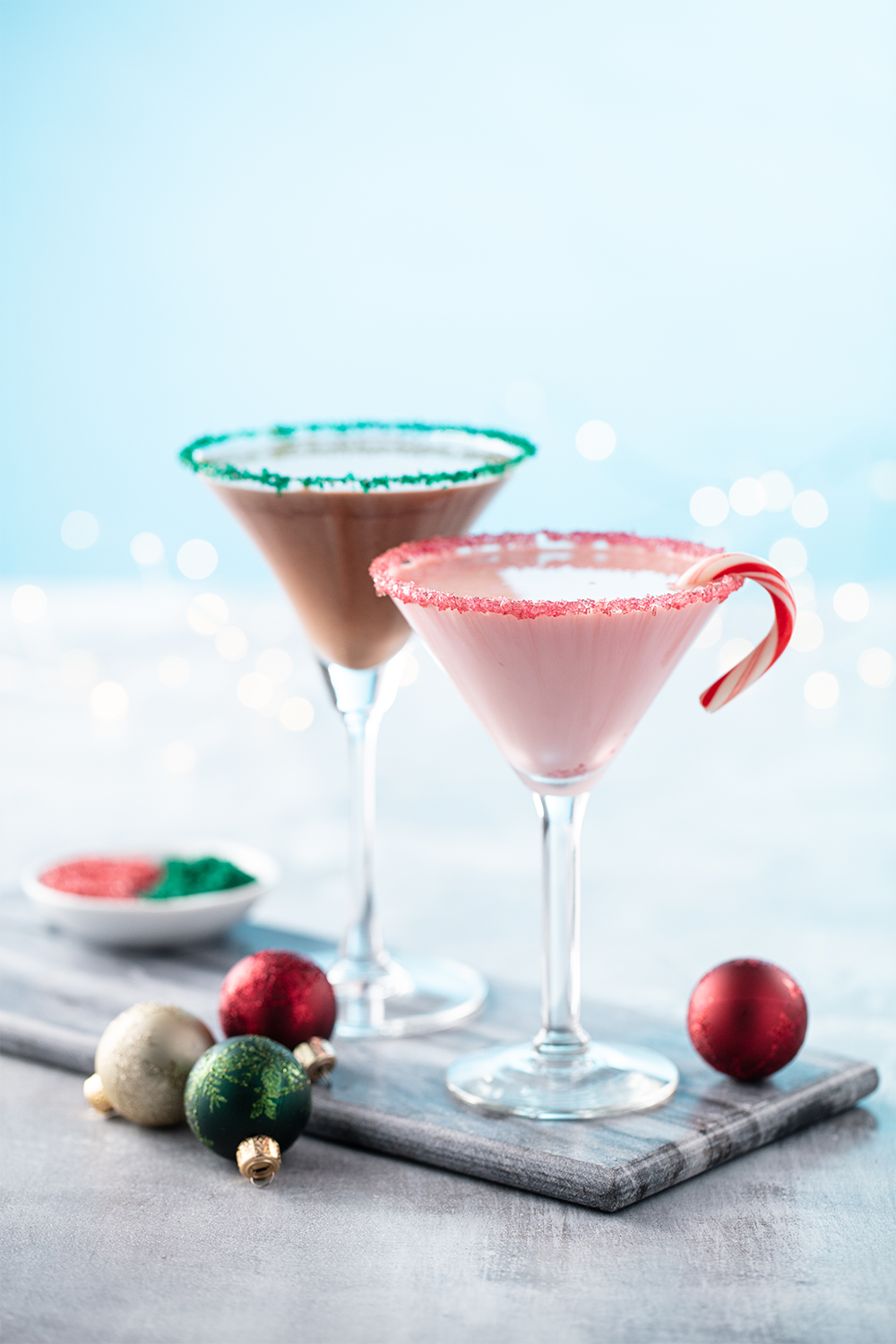 Two holiday-themed pink and brown cocktails in martini glasses with rim sugar, one garnished with a candy cane, on a wooden board with Christmas ornaments and a bowl of colored sugar, against a snowy background with bokeh lights.