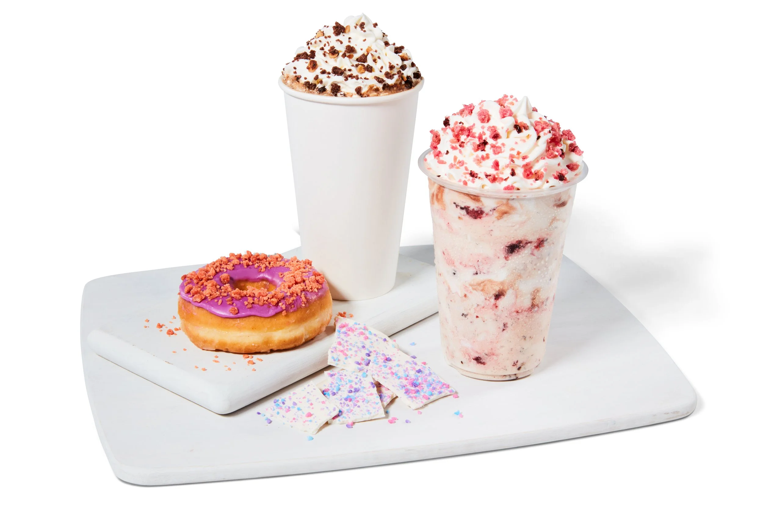 A white wooden tray with a pink glazed donut topped with pink crumbs, a white milkshake topped with whipped cream and chocolate crumbs, a berry and cream flavored milkshake topped with whipped cream and pink crumbs, and pieces of white chocolate with colorful sprinkles.