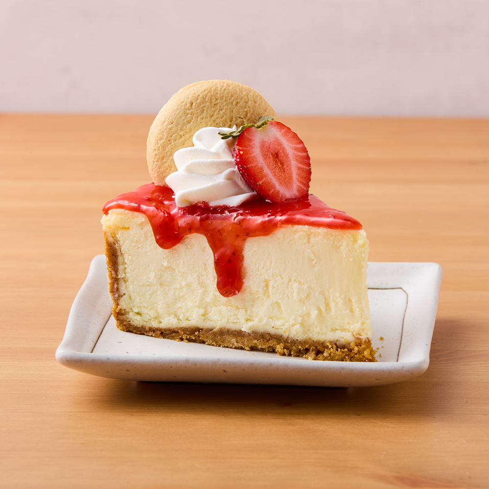 A slice of cheesecake topped with strawberry sauce, whipped cream, a strawberry half, a cookie, and a small sprig of herbs on a white rectangular plate.