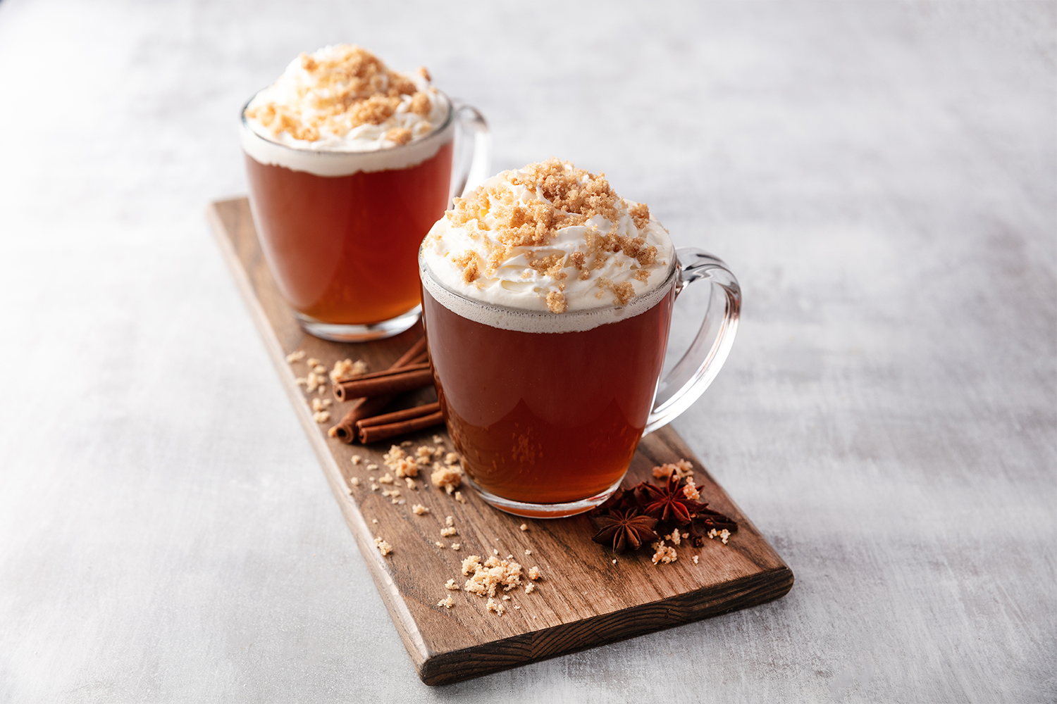 Two glass mugs of hot apple cider topped with whipped cream and crumbled streusel sit on a wooden tray with cinnamon sticks and star anise, on a light-colored surface.