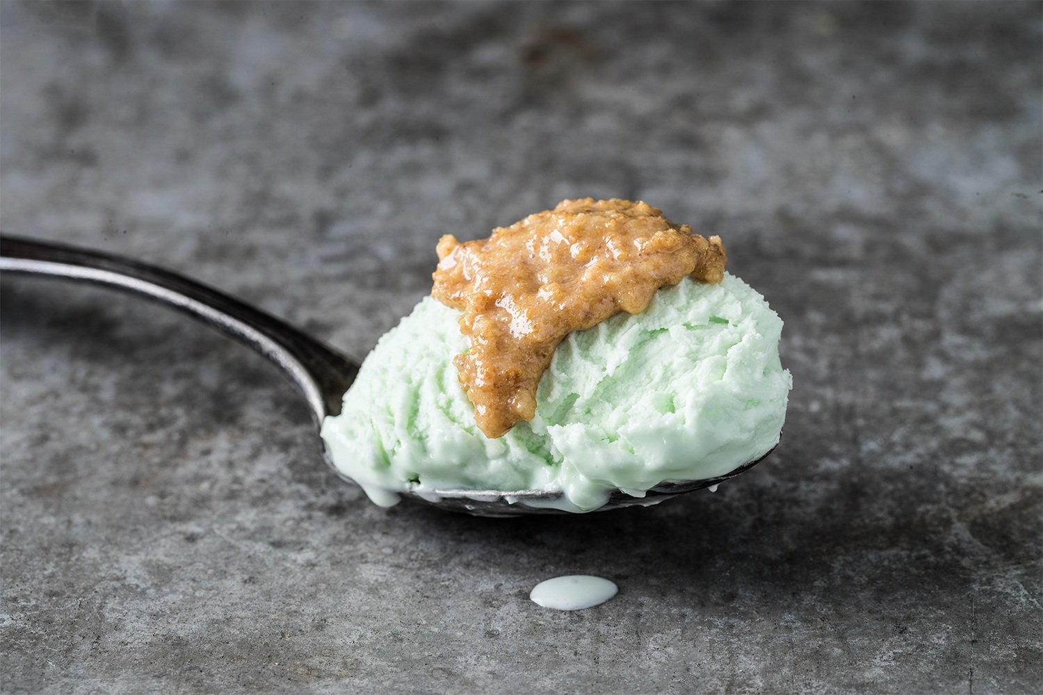 A spoonful of mint ice cream with caramel syrup on top on a gray textured surface.
