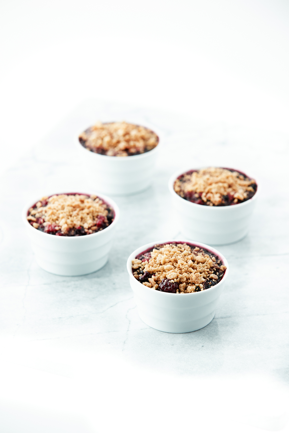 Four small white ramekins filled with berry crisps topped with streusel, placed on a white surface with a light background.