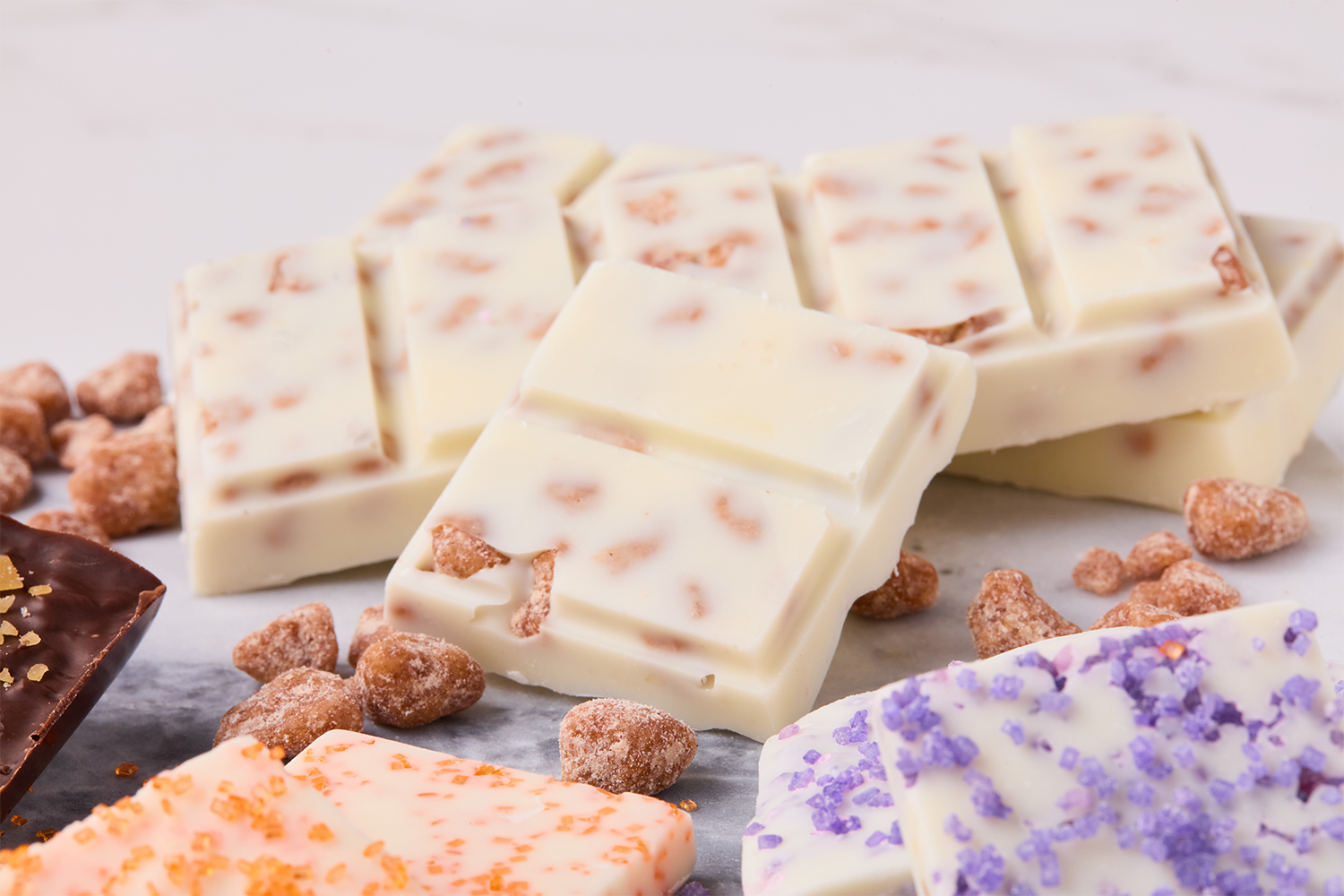 Assorted white chocolate bars with different toppings, surrounded by small sugar-coated snack pieces on a white surface.