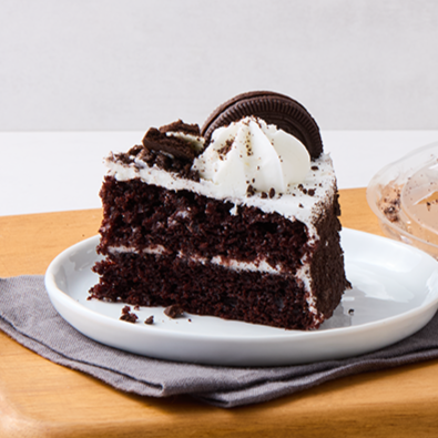 A slice of chocolate cake with white frosting, garnished with Oreo cookies and chocolate pieces on top, served on a white plate with a gray napkin underneath.