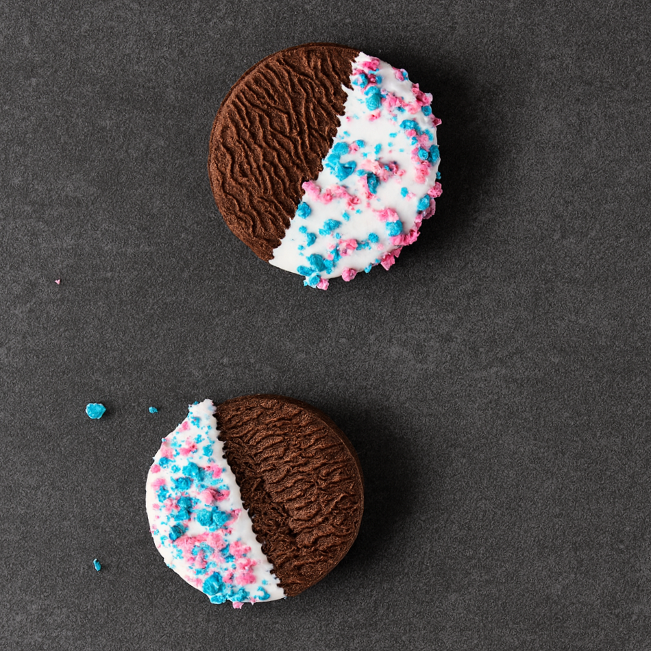 Two chocolate cookies decorated with white icing and pink and blue sprinkles, partially covered with chocolate.