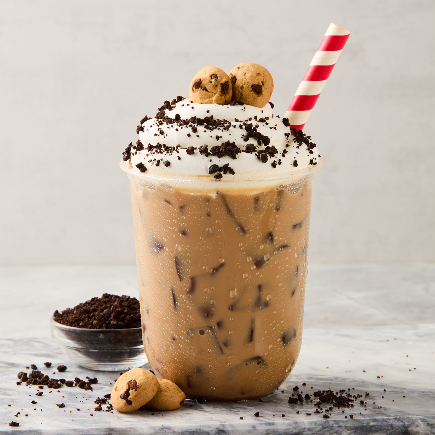 Large glass of iced coffee with whipped cream, chocolate cookie crumbs, and chocolate chip cookies on top, with a red-and-white striped straw. Small bowl of cookie crumbs and a few cookies beside the glass.