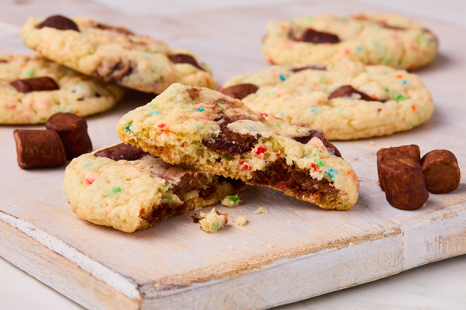 Chocolate chip cookies with colorful sprinkles and marshmallows, one broken in half, on a white wooden surface.