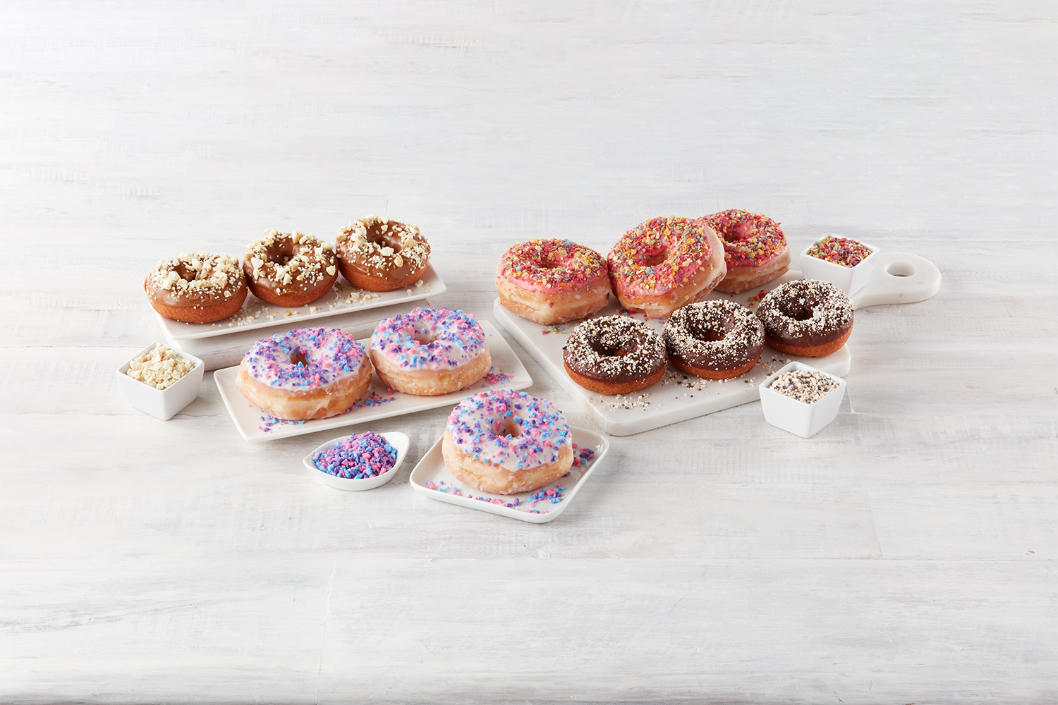 Assorted decorated donuts with sprinkles on white wooden background, displayed on white plates and small bowls.