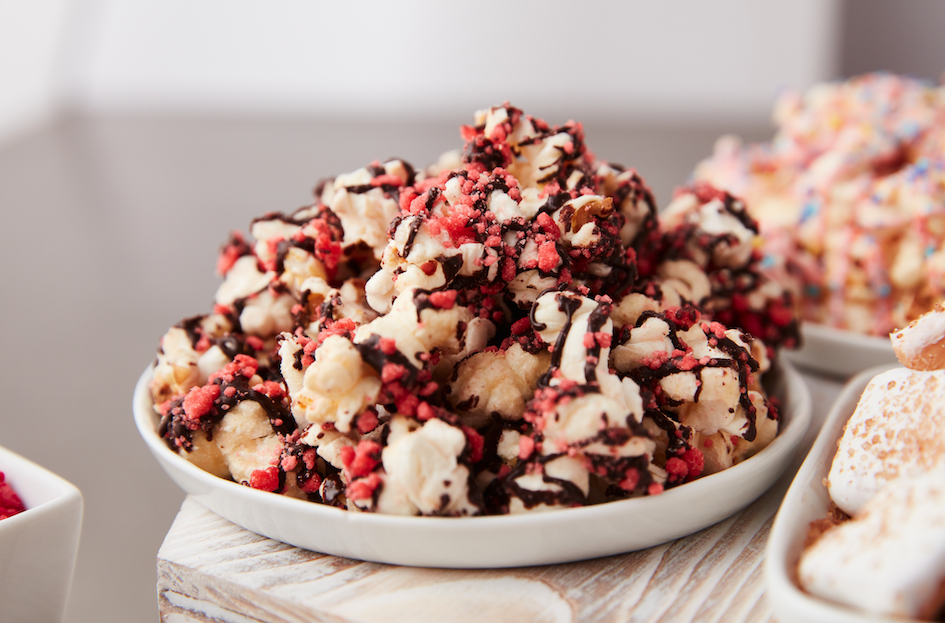 A white bowl of popcorn drizzled with chocolate and topped with pink and red crunchy candy crumbles, placed on a wooden surface.