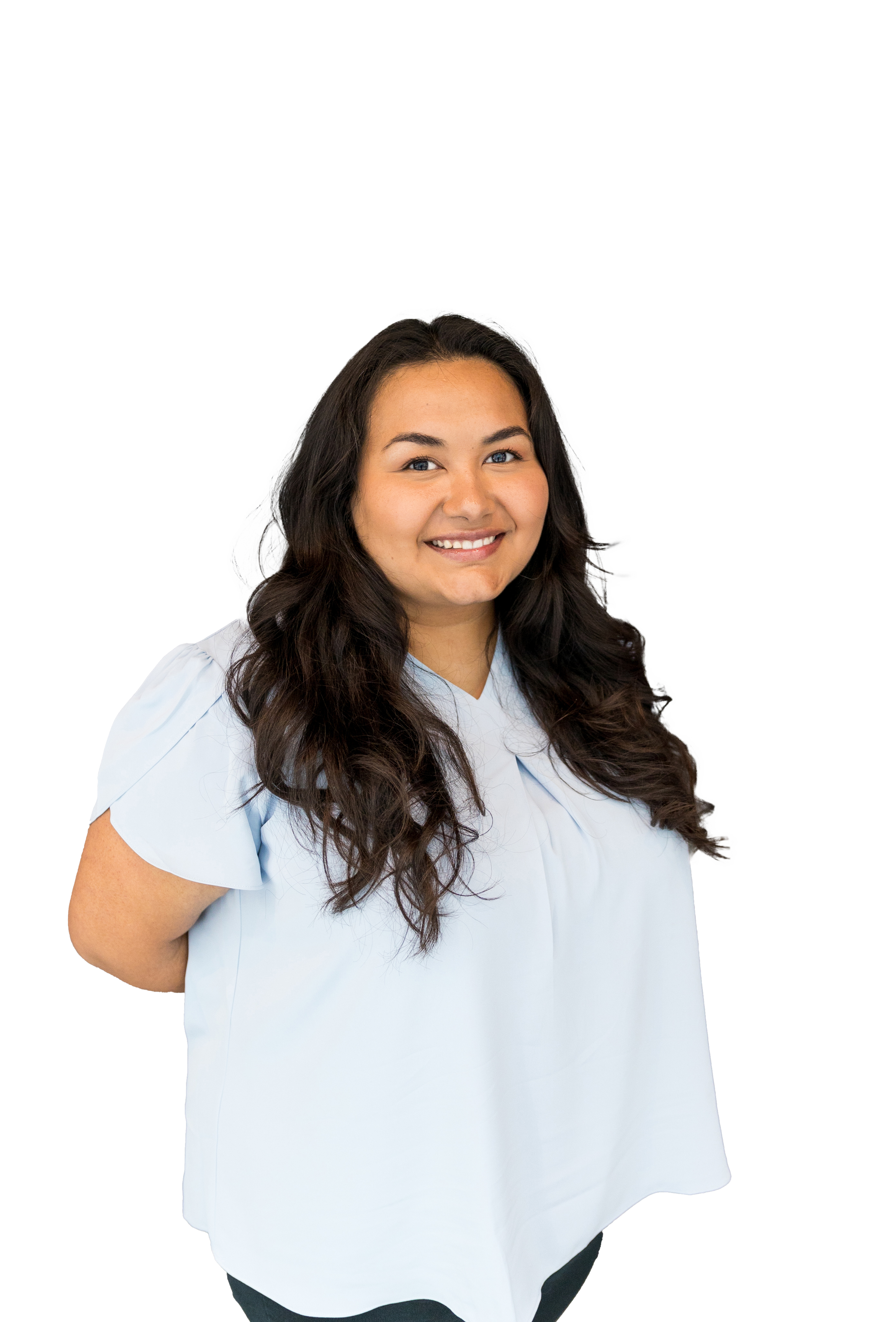 A smiling woman with long dark wavy hair wearing a light blue short-sleeved blouse standing against a transparent background.
