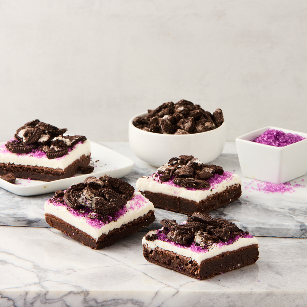 Chocolate Oreo cheesecake bars topped with crushed Oreos and pink sprinkles, with bowls of crushed Oreos and pink sugar on a marble surface.