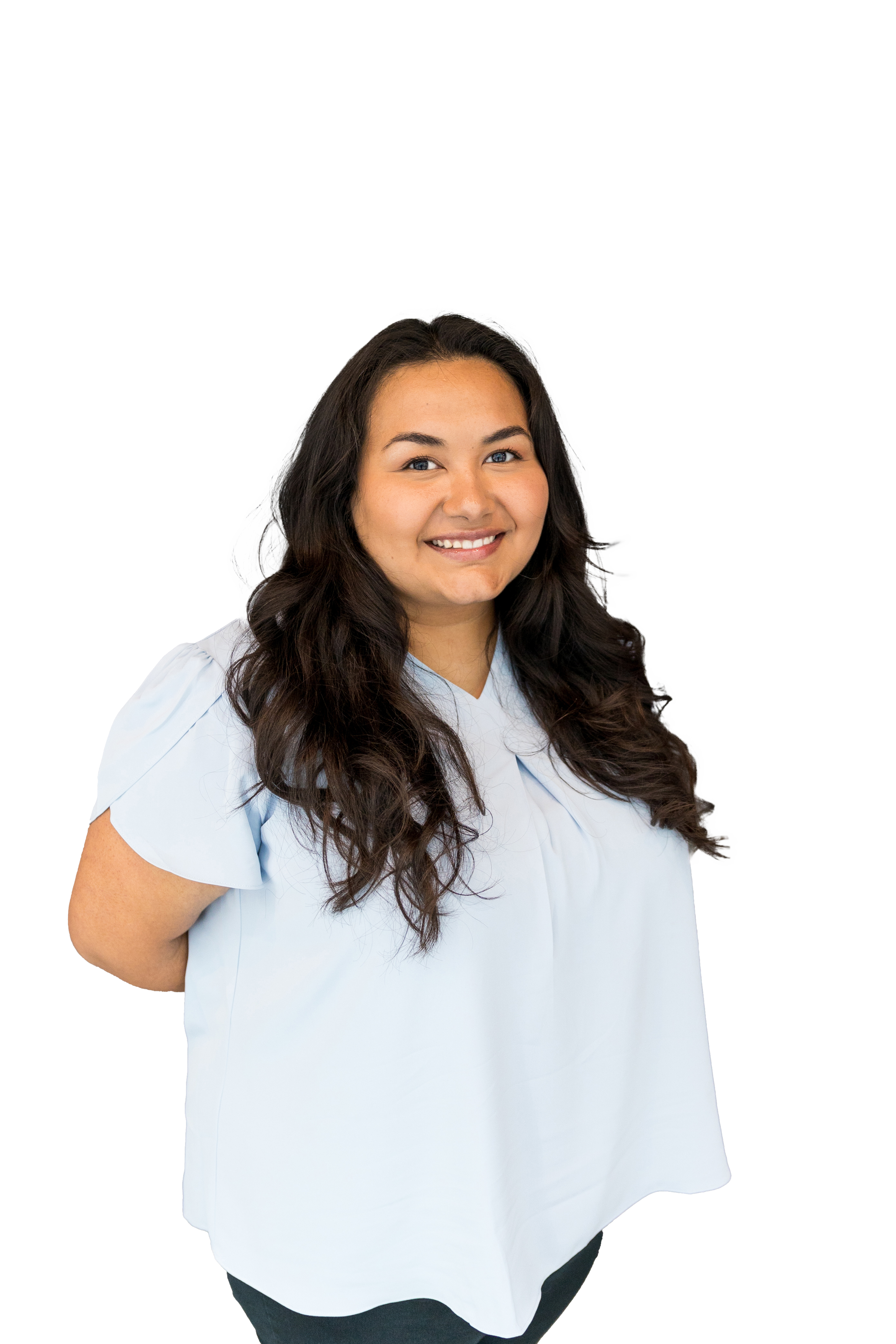 A woman with long, wavy dark hair, smiling, wearing a light blue blouse with short sleeves and black pants, standing against a white background.