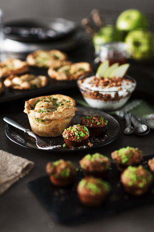 Assorted muffins and small desserts on black plates with apples and a bowl of whipped cream in the background.