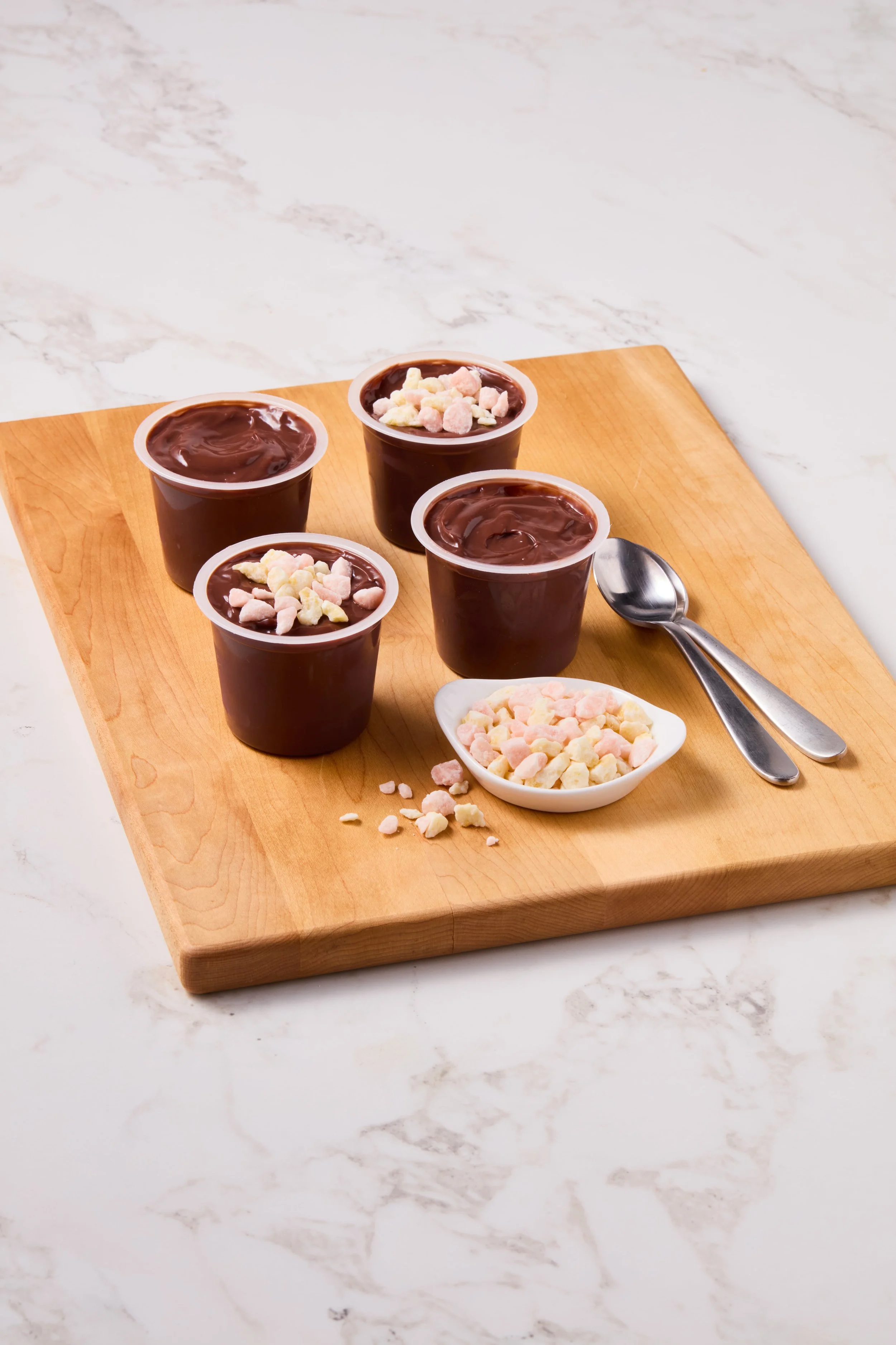 Four small cups of chocolate pudding garnished with marshmallow pieces on a wooden board, with a small spoon and a dish of marshmallow topping.