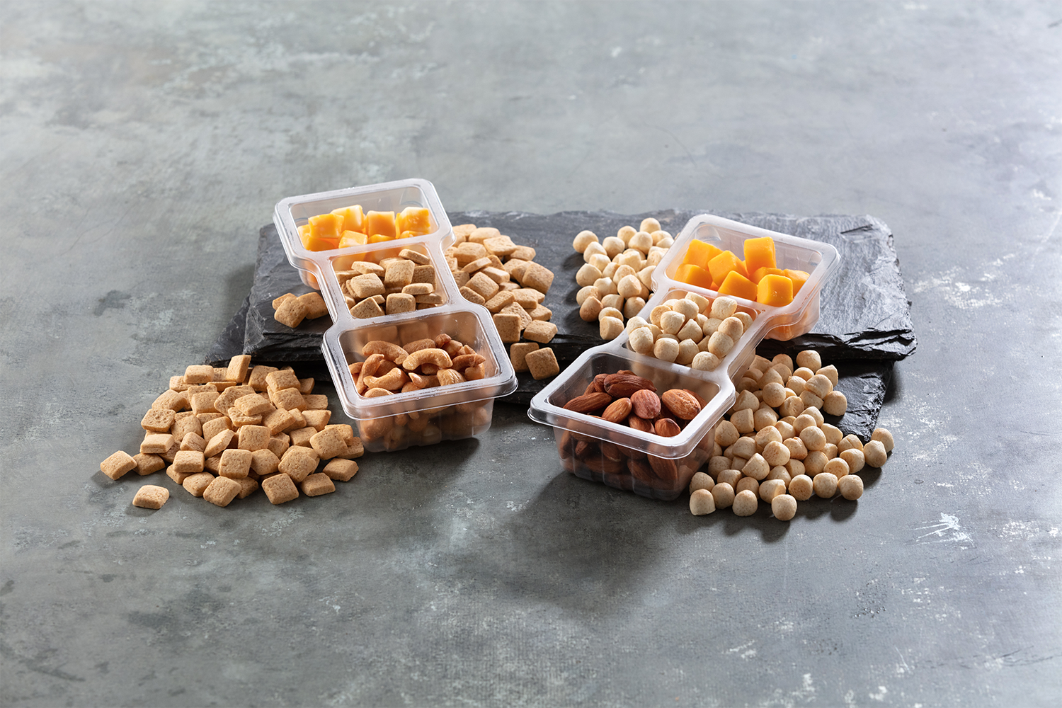 Assorted snack mix with cheese cubes, nuts, and crunchy snack squares on a dark slate platter against a gray surface.