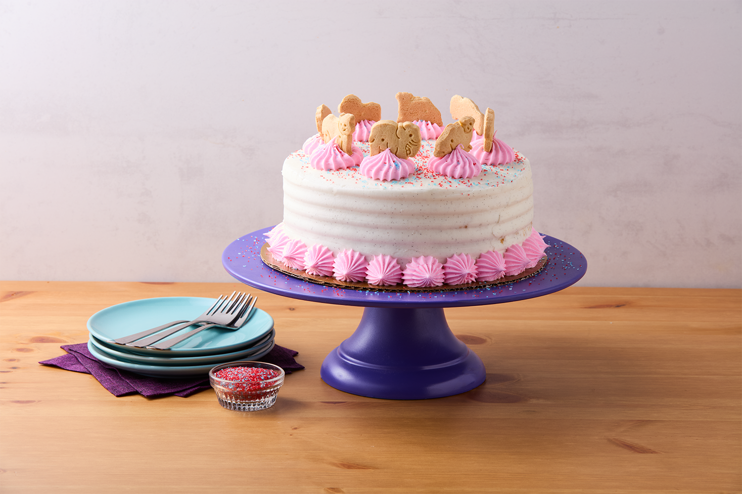 A birthday cake with white frosting, pink piped decorations, and animal-shaped cookies on top, placed on a purple cake stand. Next to the cake are stacked turquoise plates with forks, a purple napkin, and a small bowl of red sprinkles on a wooden table.