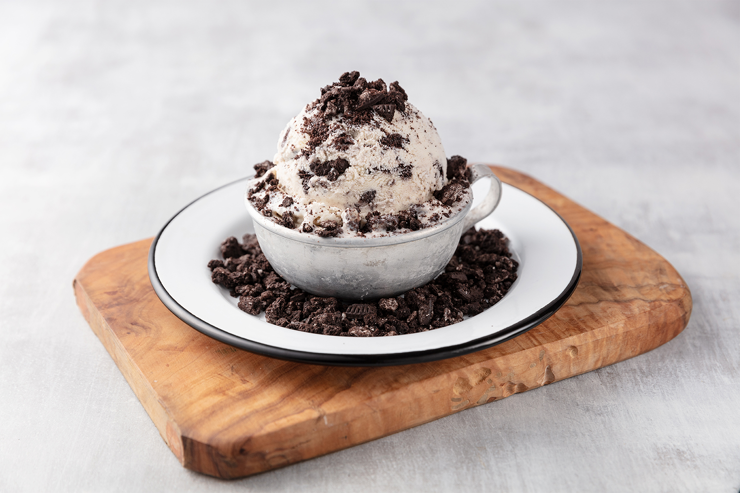 Oreo ice cream topped with crushed Oreo cookies in a gray cup on a white plate, garnished with crushed Oreo, on a wooden cutting board.