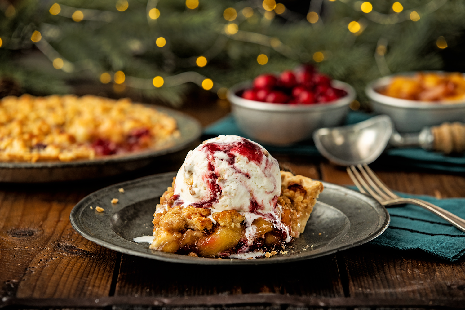A slice of cherry pie topped with vanilla ice cream on a black plate, with a whole cherry pie, bowls of cherries and fruit, and holiday decorations in the background.