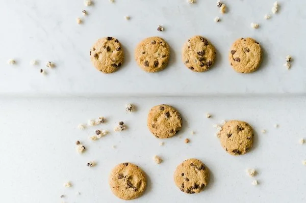 Four chocolate chip cookies on a light surface, with crumbs and four additional cookies below, some scattered crumbs around.