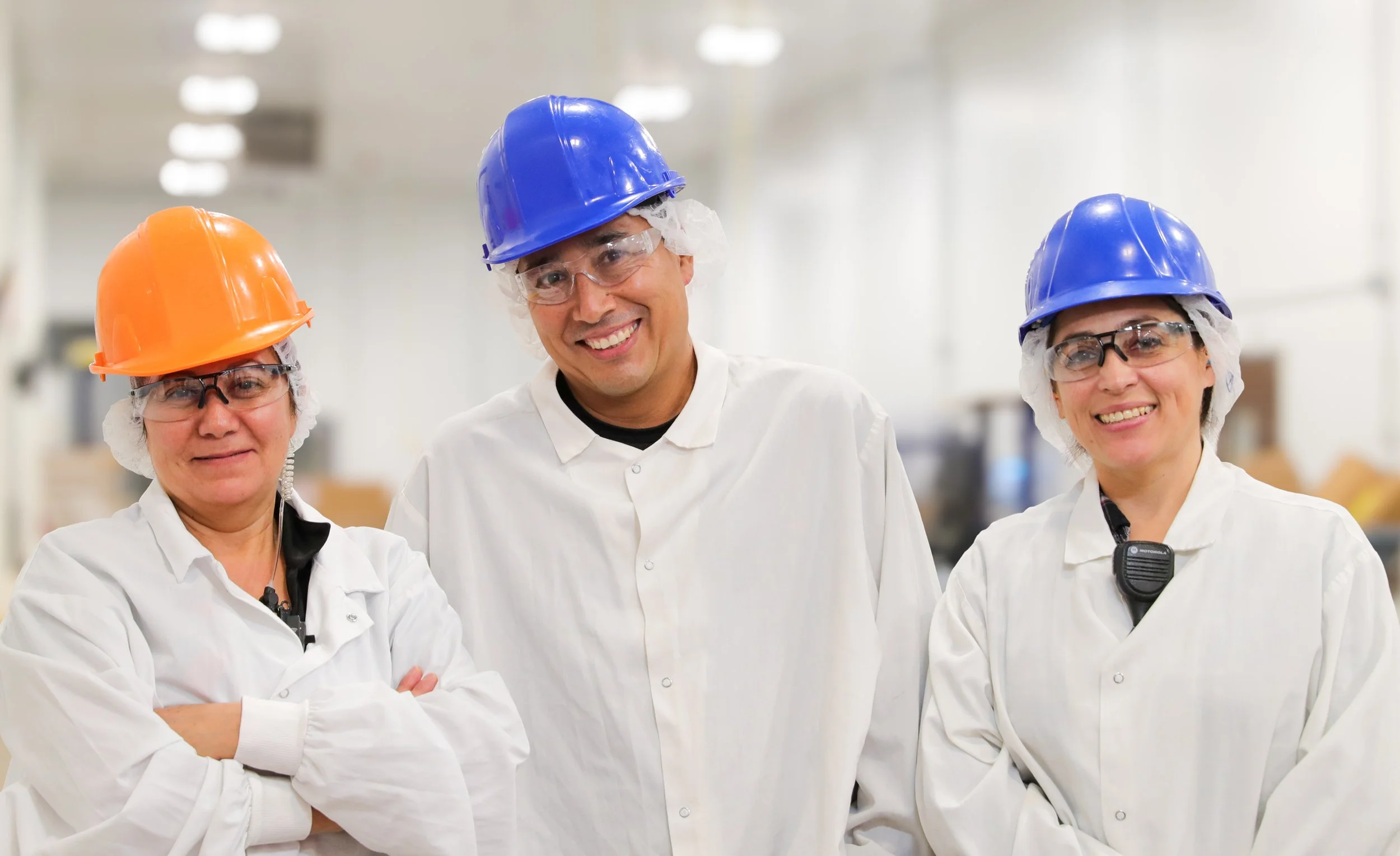 Three people in safety gear, including hard hats, safety glasses, and white lab coats, standing in an industrial or laboratory setting, smiling at the camera.
