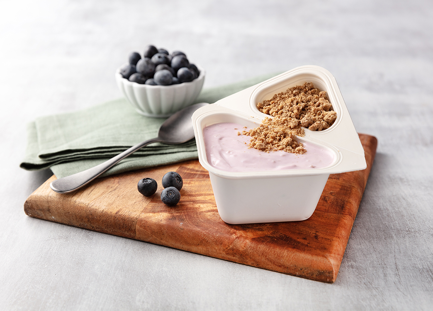 A container of strawberry yogurt with granola on top, placed on a wooden cutting board. Behind it, there is a bowl of fresh blueberries, a spoon, and a green cloth napkin. Three blueberries are on the cutting board.